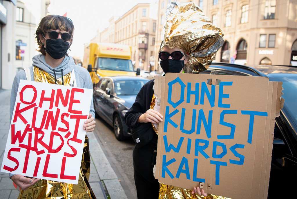 Auf der Münchner Maximilianstraße: Eine Frau und ein Mann mit Masken und Sonnenbrillen. Sie halten Demonstrationsschilder hoch, auf denen geschrieben steht: "Ohne Kunst wird's still" sowie "Ohne Kunst wird's kalt"  