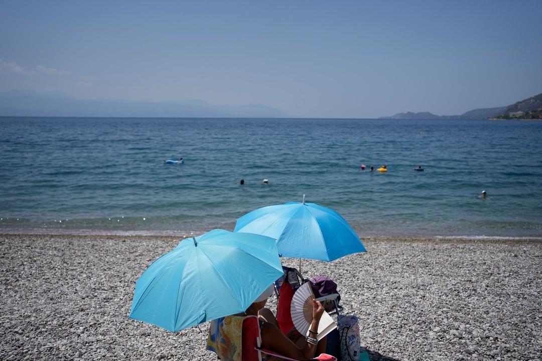 Eine Frau mit einem Handfächer sitzt unter einem Sonnenschirm in Griechenland am Strand. (Symbolbild)