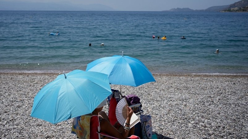 Eine Frau mit einem Handfächer sitzt unter einem Sonnenschirm in Griechenland am Strand. (Symbolbild) | Bild: dpa-Bildfunk/Thanassis Stavrakis Eine Frau mit einem Handfächer sitzt unter einem Sonnenschirm in Griechenland am Strand. (Symbolbild)