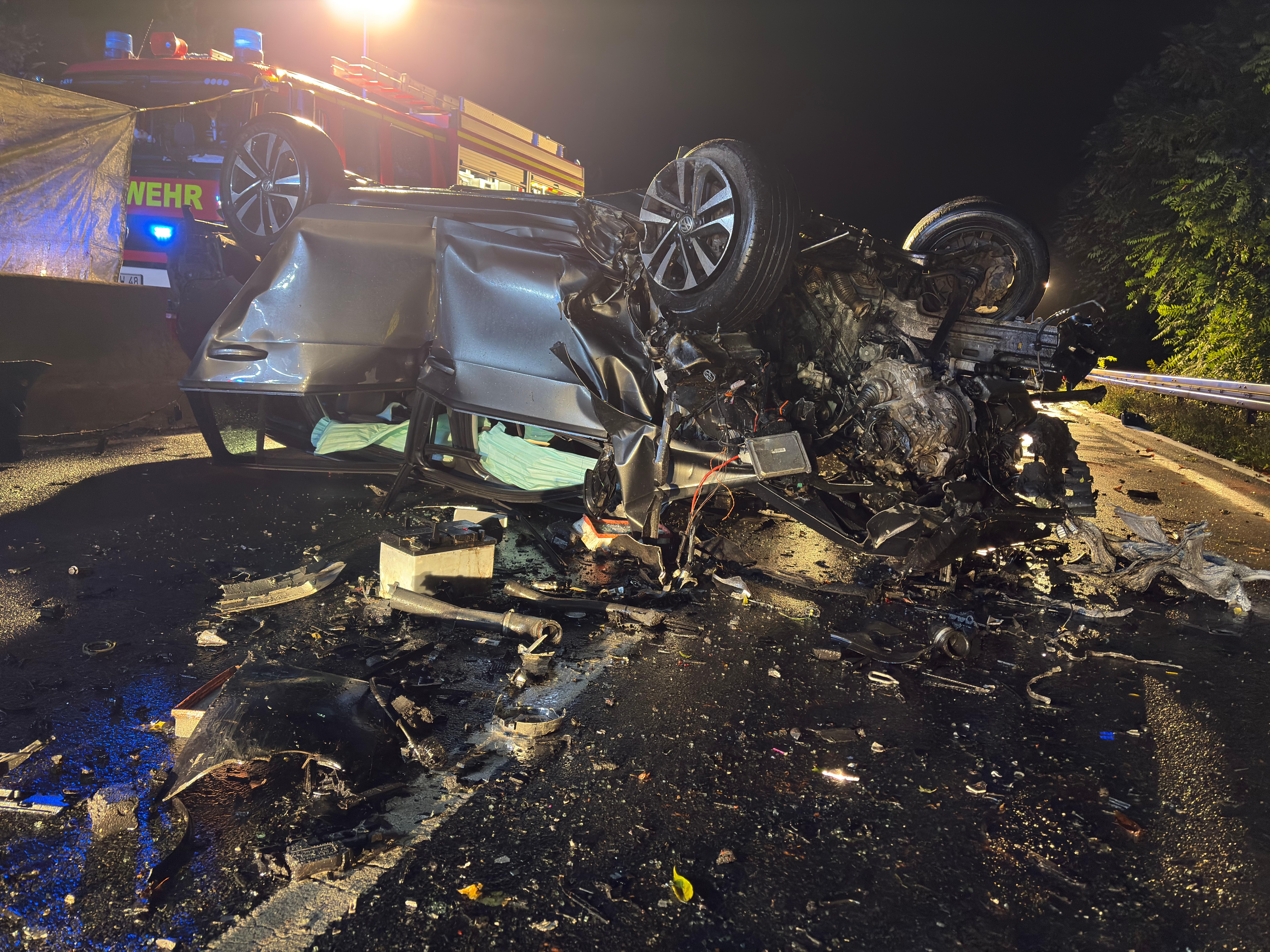 Ein völlig zerstörtes Autowrack liegt auf dem Dach. Im Hintergrund ist ein Feuerwehrfahrzeug zu sehen. 