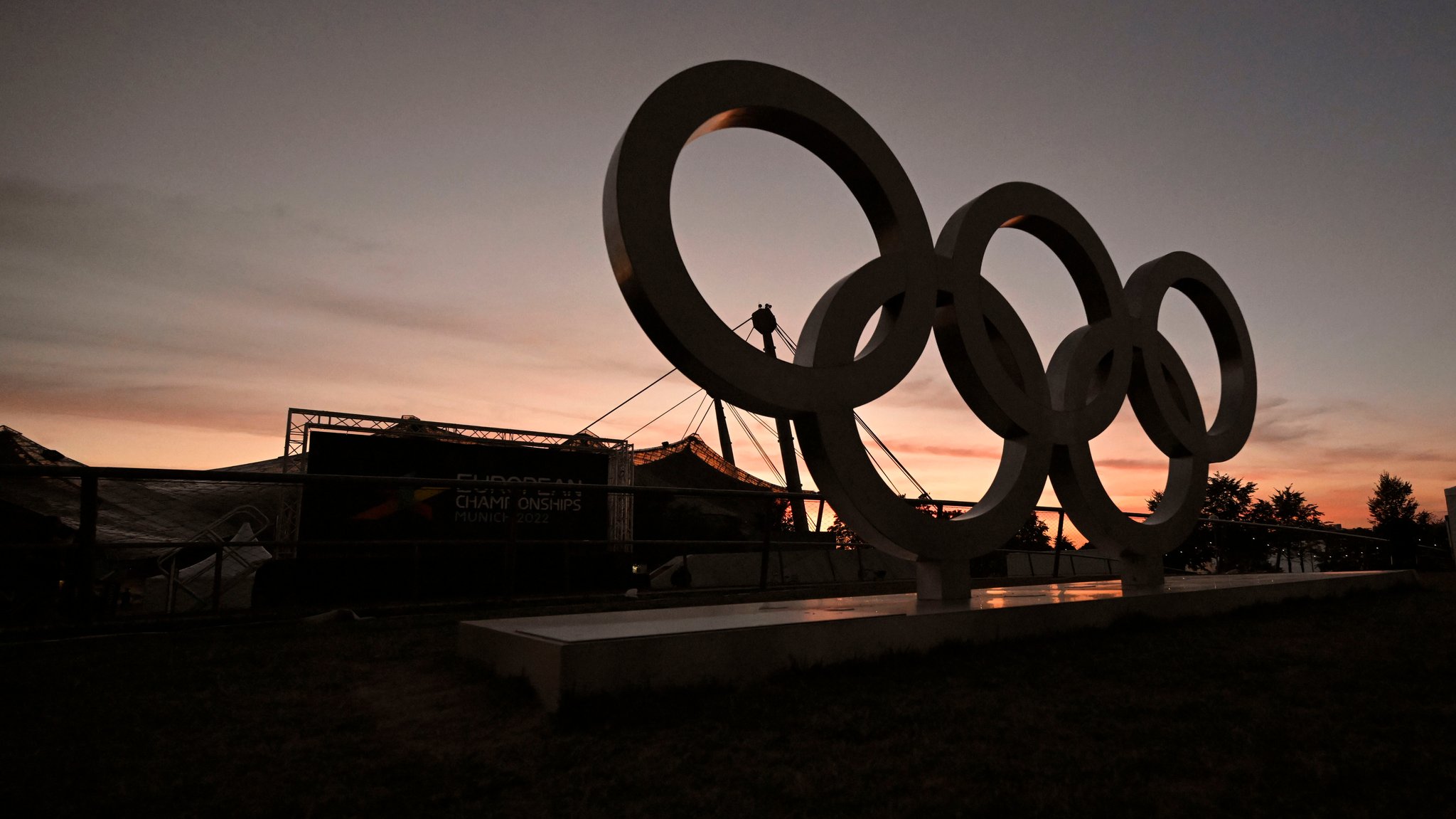 Die olympischen Ringe stehen 2022 während der European Championships in München im Olympiapark. Im Hintergrund sieht man die Sportstätten und einen Sonnenuntergang | Bild: picture alliance / M.i.S. | Bernd Feil Die olympischen Ringe stehen 2022 während der European Championships in München im Olympiapark. Im Hintergrund sieht man die Sportstätten und einen Sonnenuntergang