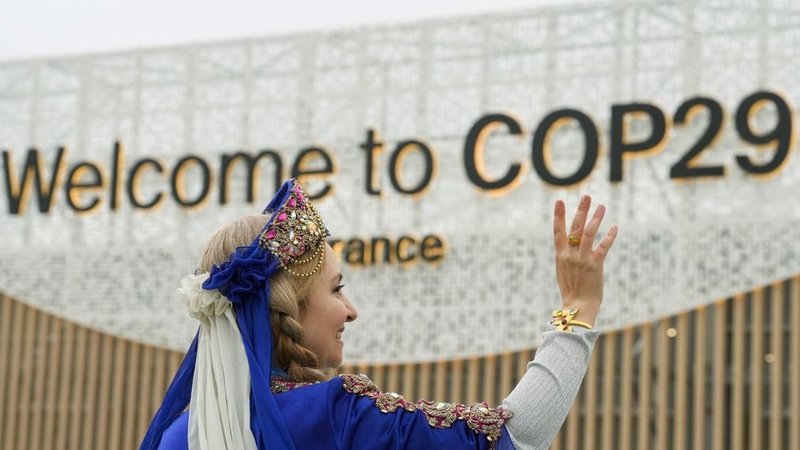 dpatopbilder - 12.11.2024, Aserbaidschan, Baku: Eine Frau in Tracht winkt vor dem Eingang zum Tagungsort des UN-Klimagipfels COP29. Foto: Peter Dejong/AP/dpa +++ dpa-Bildfunk +++ | Bild: dpa-Bildfunk/Peter Dejong dpatopbilder - 12.11.2024, Aserbaidschan, Baku: Eine Frau in Tracht winkt vor dem Eingang zum Tagungsort des UN-Klimagipfels COP29. Foto: Peter Dejong/AP/dpa +++ dpa-Bildfunk +++