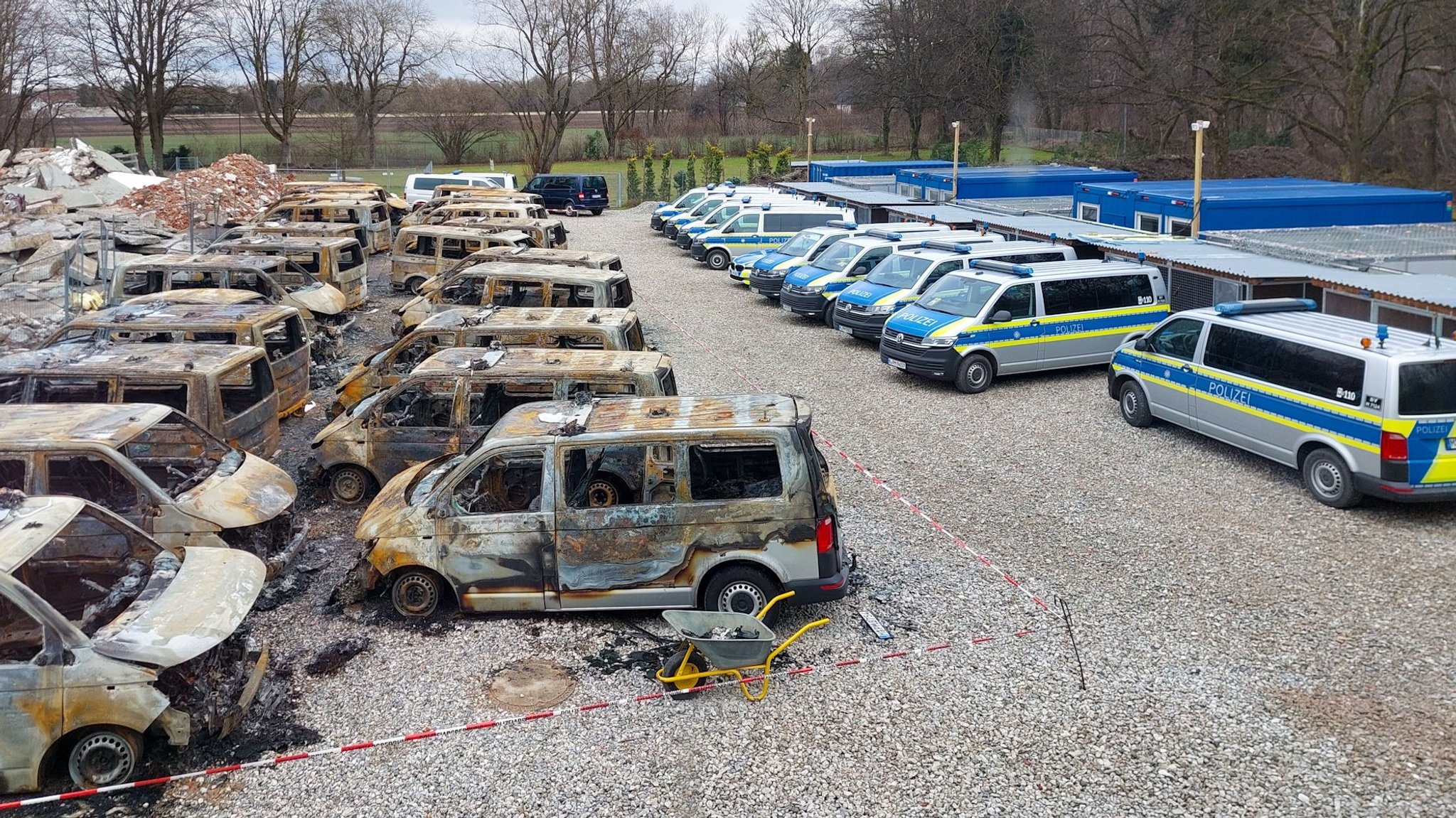 Blick aus der Vogelperspektive auf die 23 ausgebrannten Fahrzeuge auf dem Parkplatz der Münchner Polizeihundestaffel. Daneben stehen funktionstüchtige Polizeifahrzeuge. Vor knapp einer Woche sind bei einem Brandanschlag bei der Münchner Polizeihundestaffel 23 Fahrzeuge zerstört worden. 