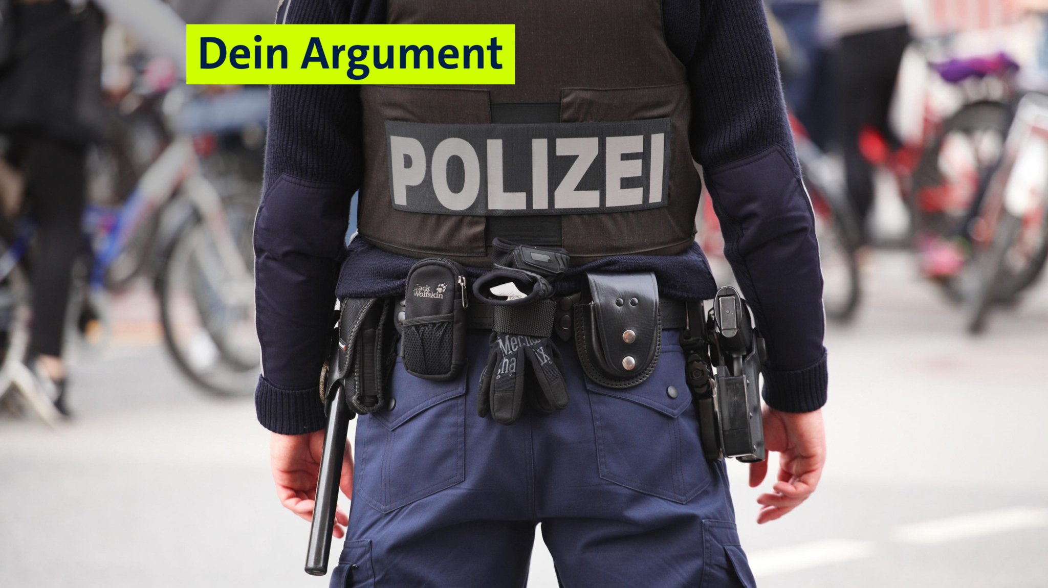 Symbolbild: Ein Polizist steht einer Straße. Er trägt eine Schutzweste, seine Pistole, einen Schlagstock, am Gürtel ein paar Handschuhe, ein Lederetui mit Handschellen und weitere Ausrüstung.
