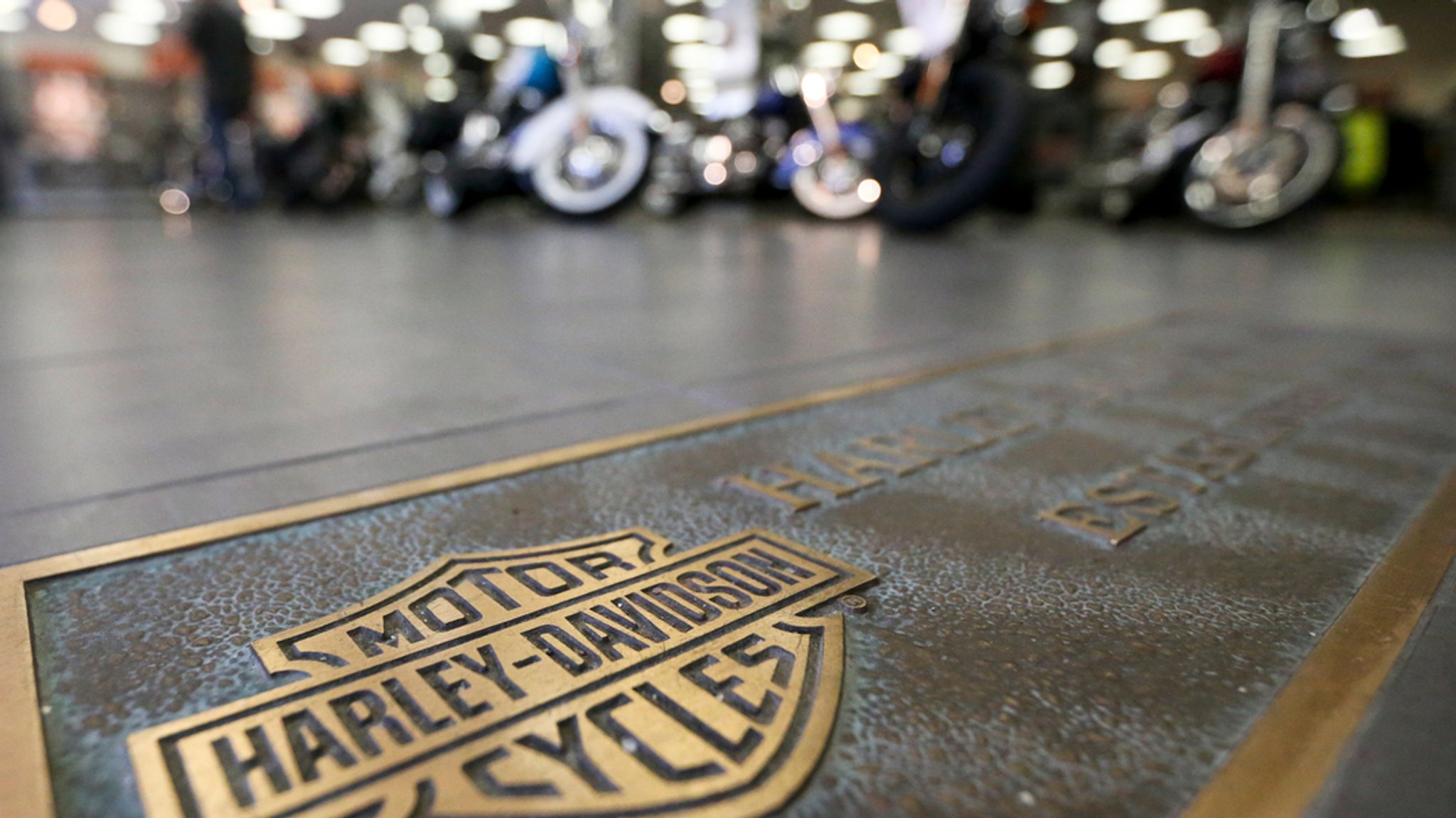 ARCHIV - 26.04.2017, USA, Glenshaw: Motorräder stehen in Reihen und ein bronzenes Emblem des Motorrad-Herstellers Harley-Davidson ist auf dem Boden eines Ausstellungsraums eines Harley-Davidson-Händlers angebracht. Der US-Motorradbauer Harley-Davidson will rechtlich gegen höhere EU-Zölle vorgehen. 