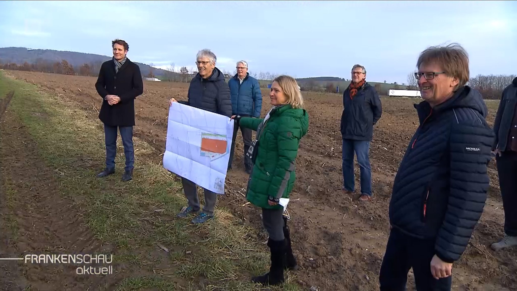 Sechs Personen stehen auf einem Acker und halten einen Bauplan in Händen.