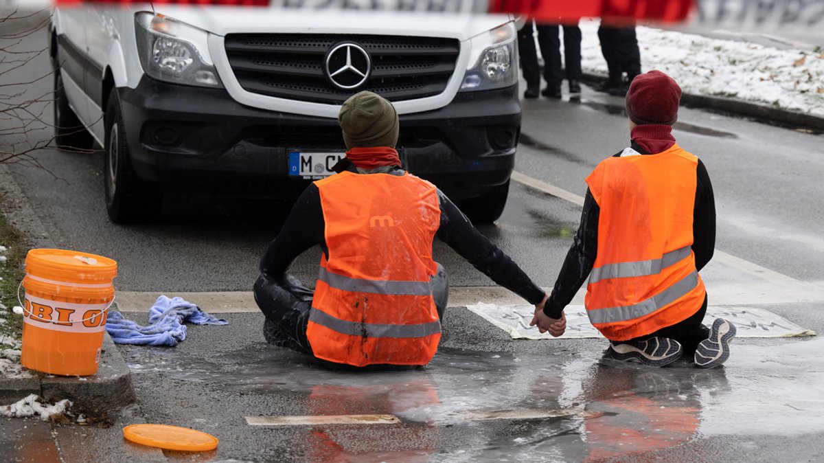 Zwei Klimaaktivisten nehmen an einer Protestaktion der Gruppe "Letzte Generation" teil, indem sie trotz Regen versuchen, sich auf einer Straße festzukleben.