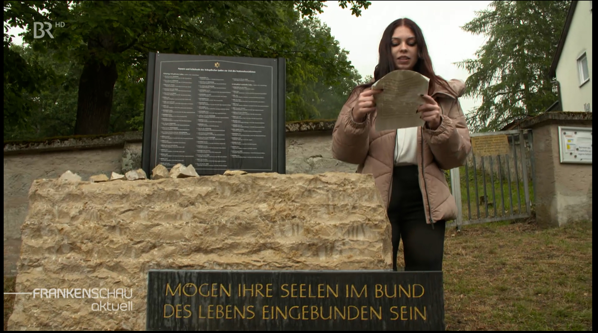 Eine Jugendliche liest neben einem Gedenkstein einen Text von einem Blatt Papier ab