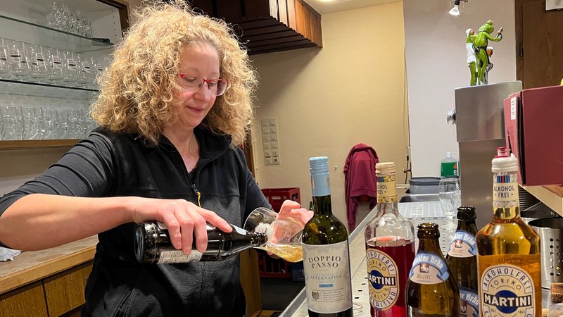 Eine Frau schenkt Bier aus einer Flasche hinter der Bar in ein Pilsglas ein. Im Vordergrund stehen Getränkeflaschen mit der Aufschrift "Alkoholfrei".