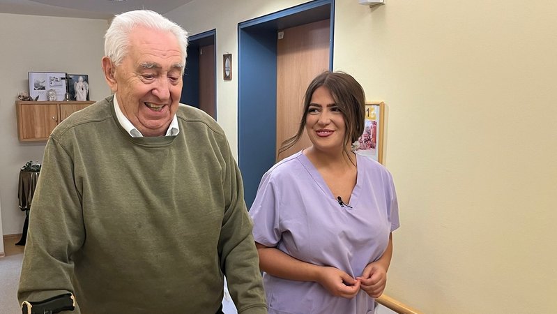 Ein Senior und eine Auszubildende laufen in einem Seniorenheim einen Gang entlang. | Bild: BR/Richard Padberg Ein Senior und eine Auszubildende laufen in einem Seniorenheim einen Gang entlang.