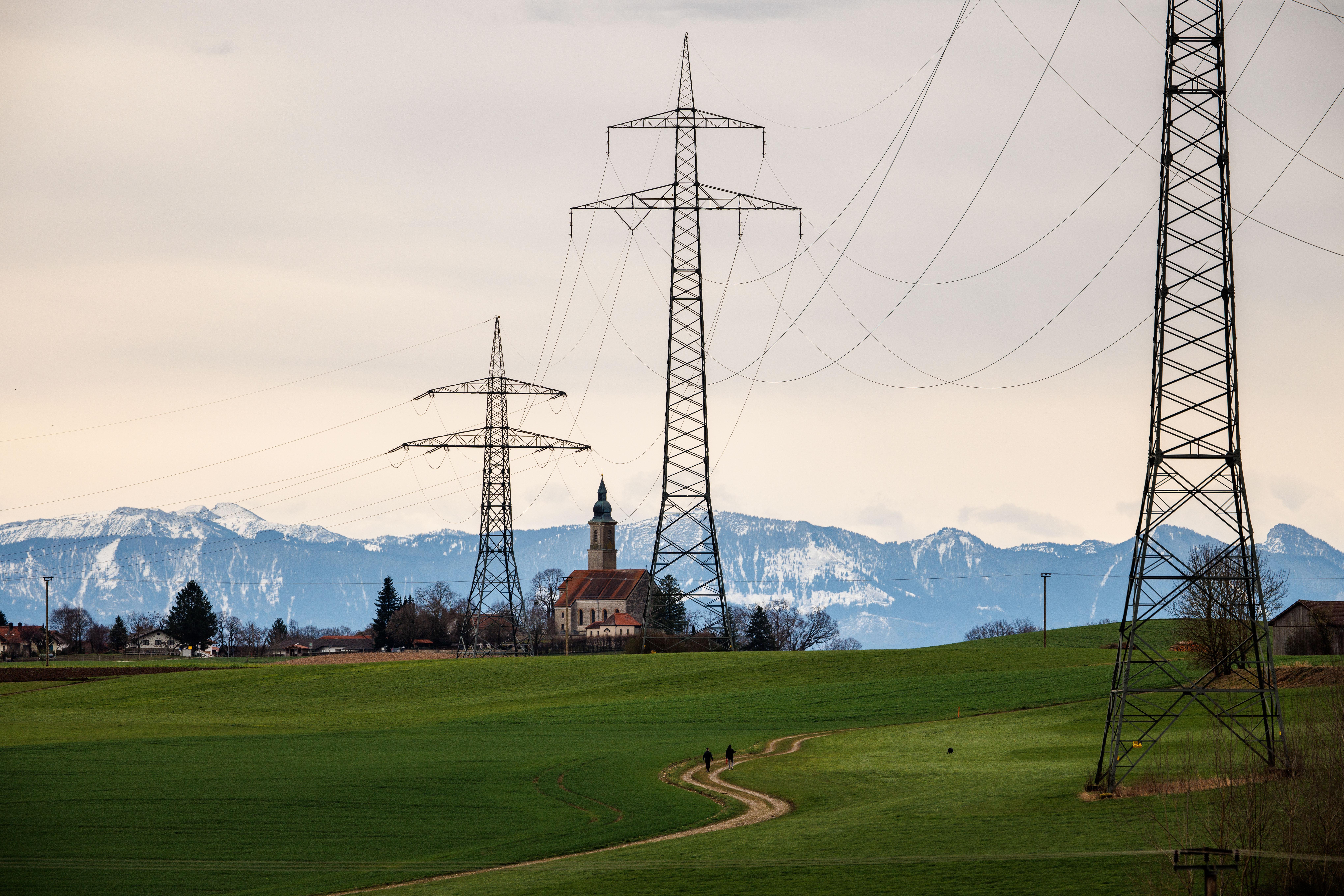 Die Kirche St. Michael im Ort Alxing zwischen den Masten einer Hochspannungsleitung vor der Alpenkette