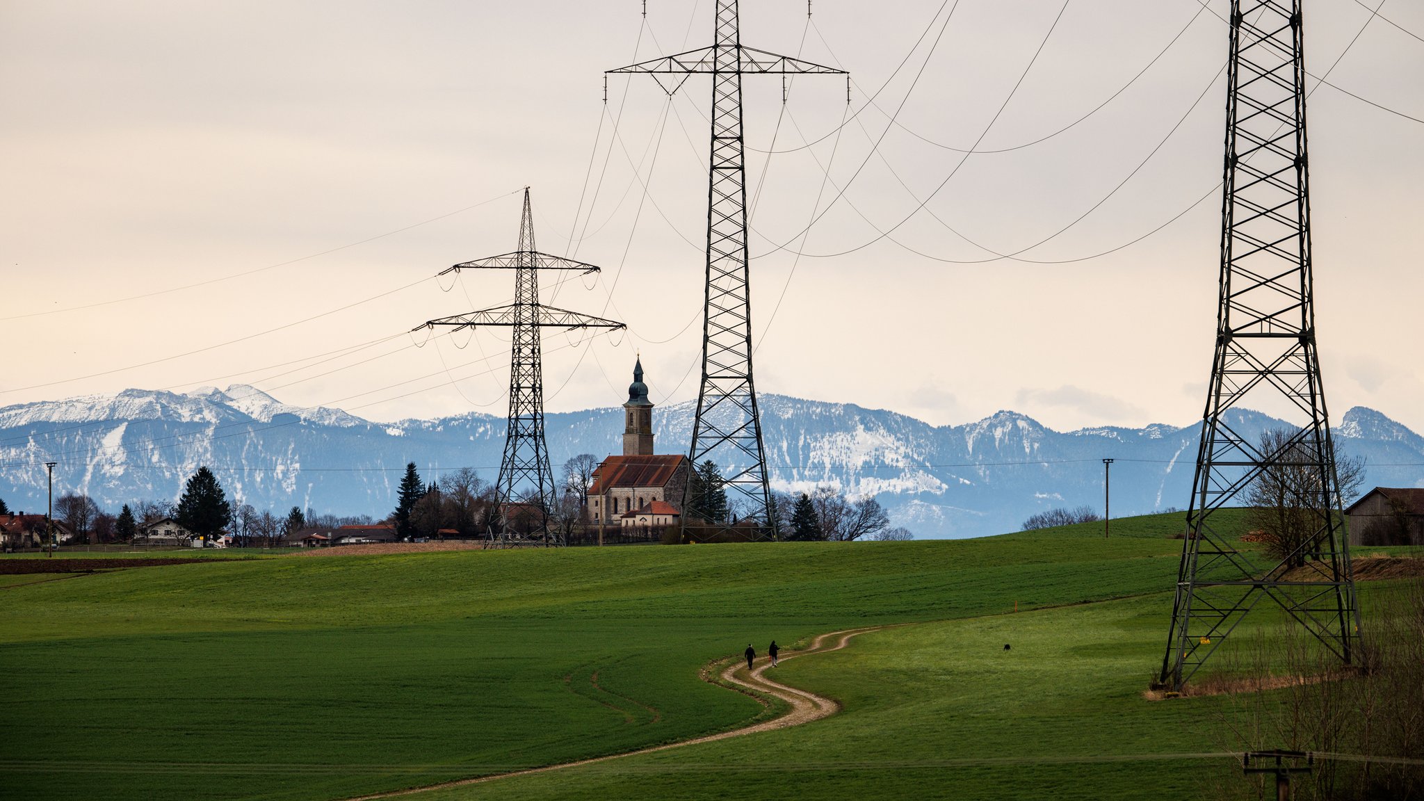 Die Kirche St. Michael im Ort Alxing zwischen den Masten einer Hochspannungsleitung vor der Alpenkette