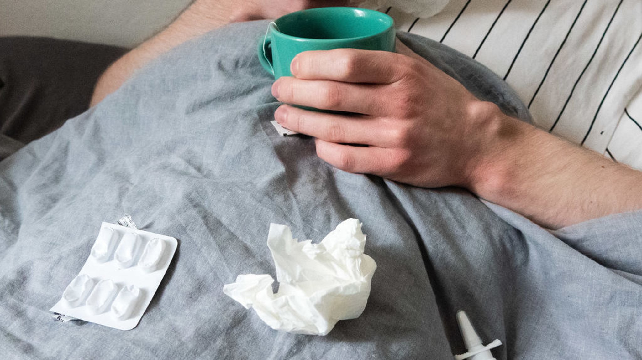 Ein Mann liegt mit Taschentüchern, Teetasse, Nasenspray und Tabletten im Bett