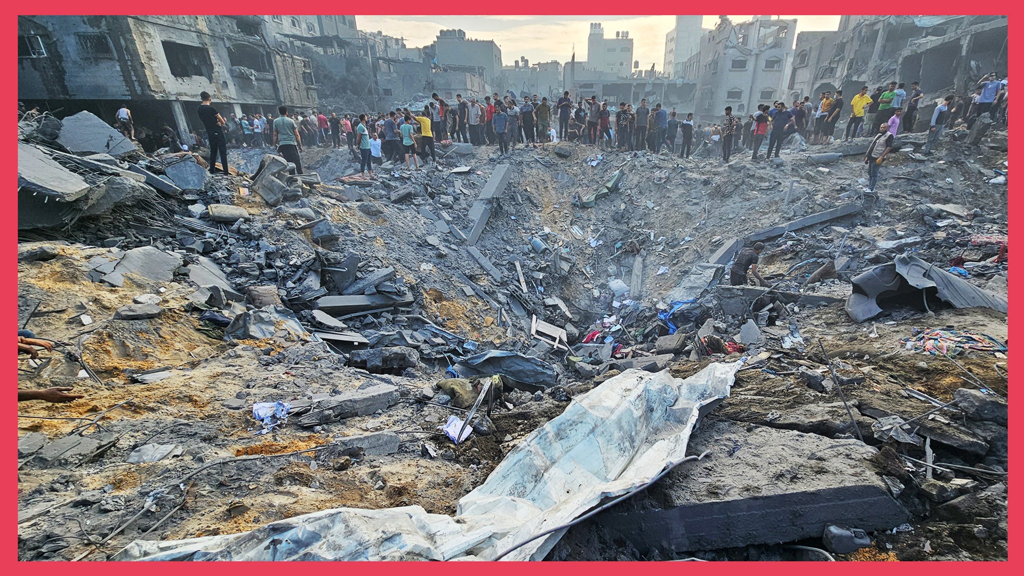 Ein Krater mit Trümmerteilen, im Hintergrund Menschen und eine zerstörte Skyline von Gaza-Stadt.