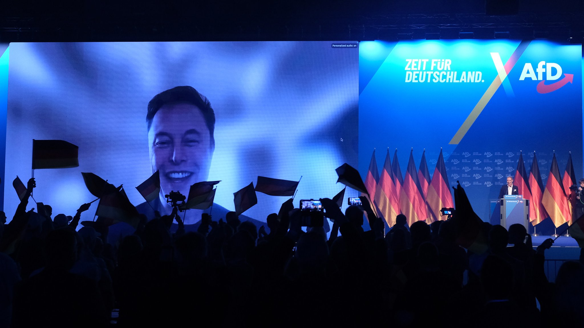 Auf dem Bild sieht man den AfD-Parteitag in Halle an der Saale. Auf eine Leinwand ist Elon Musk projiziert, der sich dem Parteitag per Videocall zugeschaltet hat. Rechts daneben steht die AfD-Vorsitzende Alice Weidel auf einer Bühne. Im Vordergrund schwenken Menschen Deutschlandflaggen.