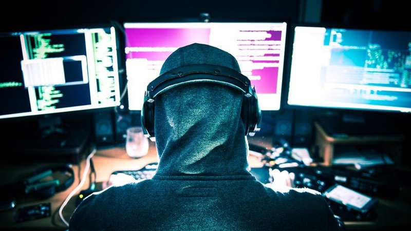 Eine Person mit Kapuzenpullover und Kopfhörern sitzt vor mehreren Monitoren, die mit Code und Daten gefüllt sind. (Symbolbild) | Bild: stock.adobe.com/peterzayda Eine Person mit Kapuzenpullover und Kopfhörern sitzt vor mehreren Monitoren, die mit Code und Daten gefüllt sind. (Symbolbild)