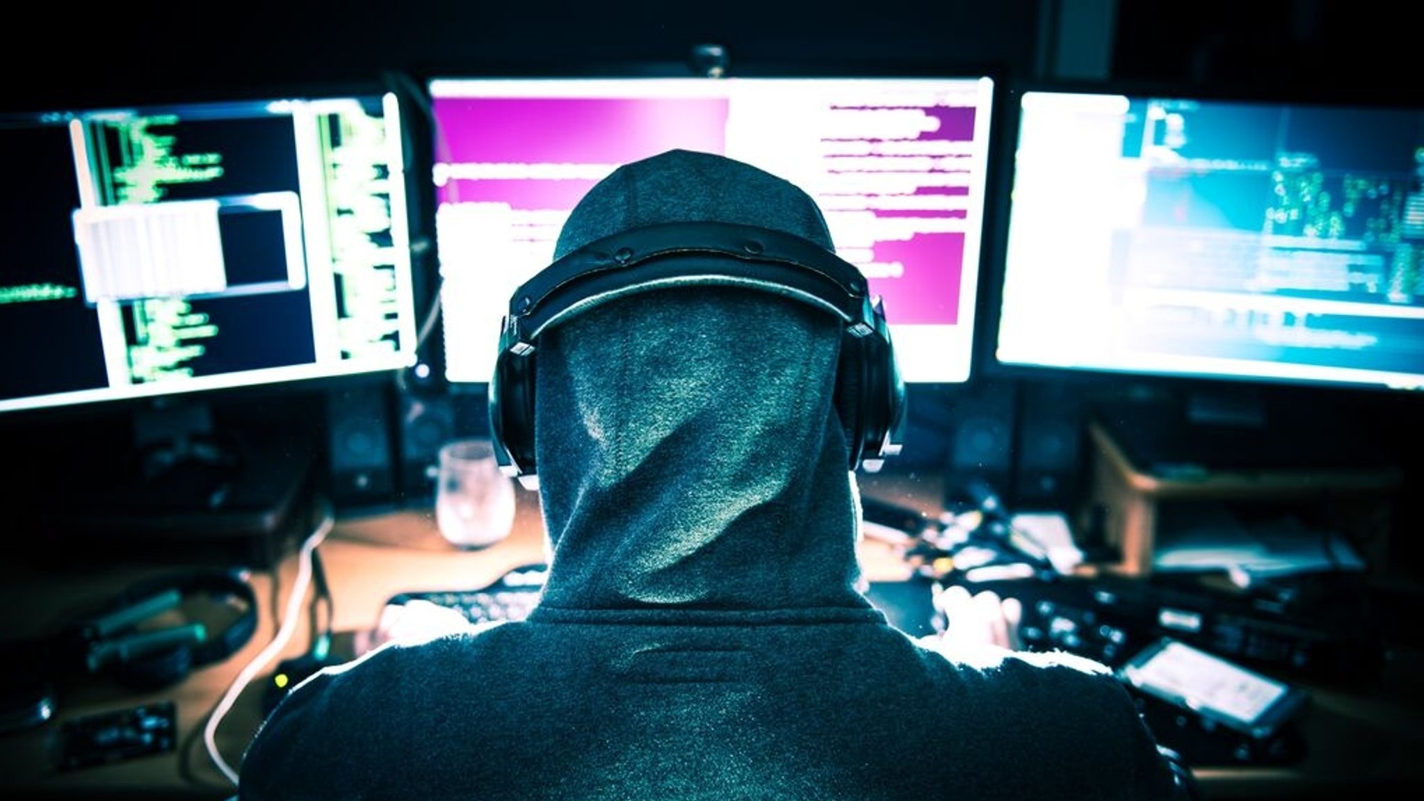 Eine Person mit Kapuzenpullover und Kopfhörern sitzt vor mehreren Monitoren, die mit Code und Daten gefüllt sind. (Symbolbild)