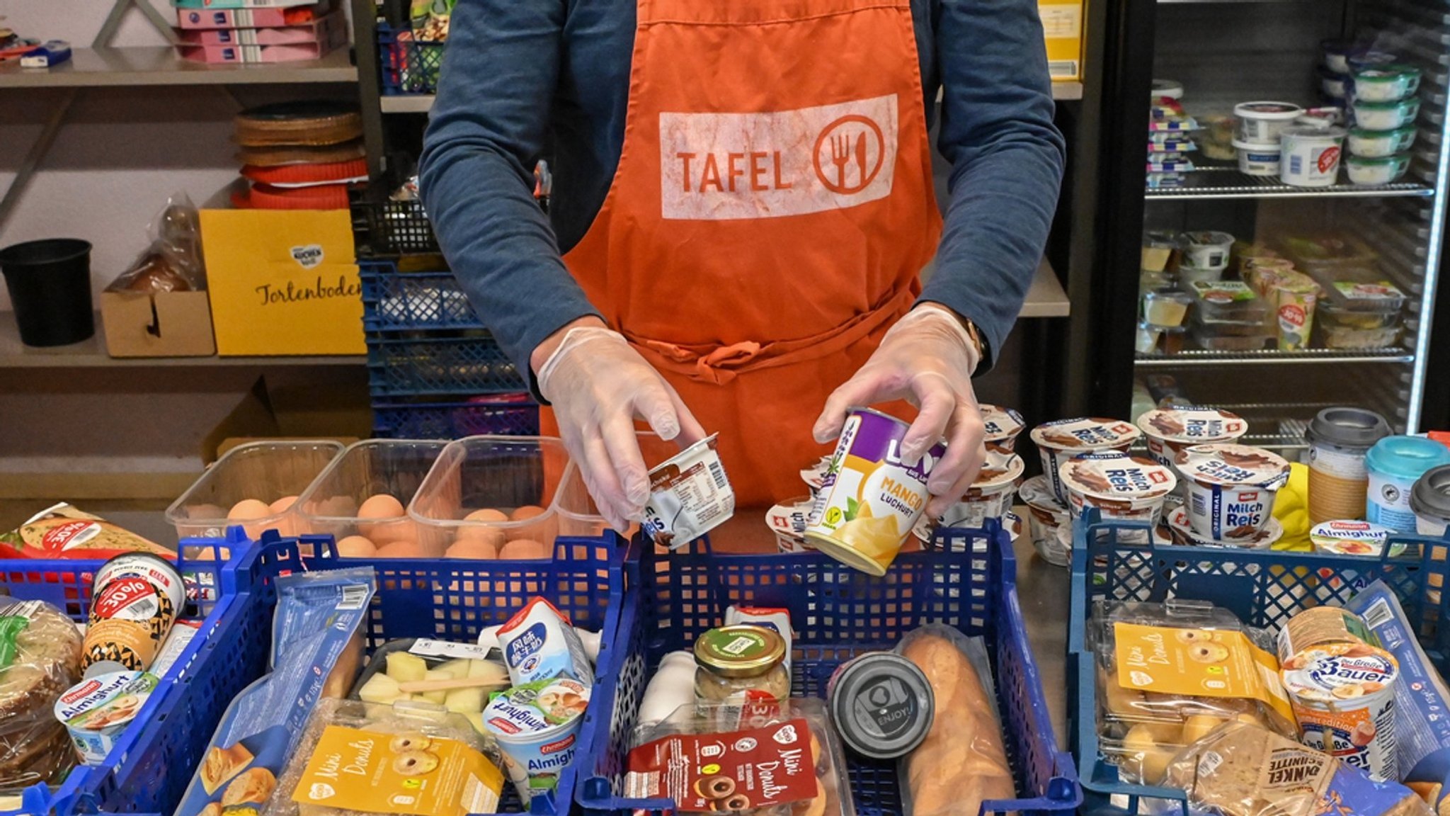 Eine Person, die eine orangene Schürze mit der Aufschrift "Tafel" trägt, steht hinter gefüllten Lebensmittelkisten. (Symbolbild)