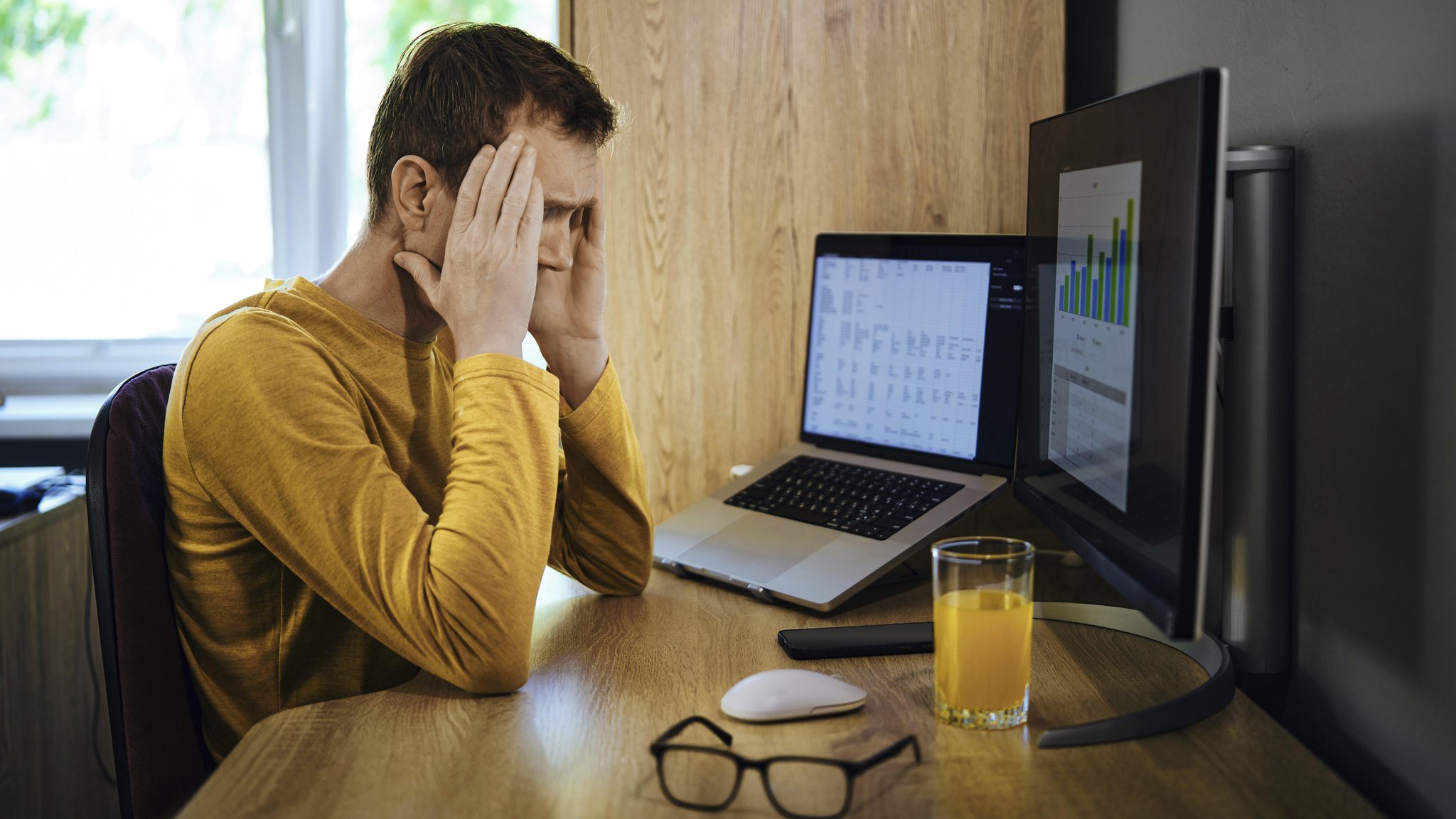 Symbolbild: Mann sitzt an einem Schreibtisch mit zwei Computerbildschirmen und hält sich frustriert den Kopf.