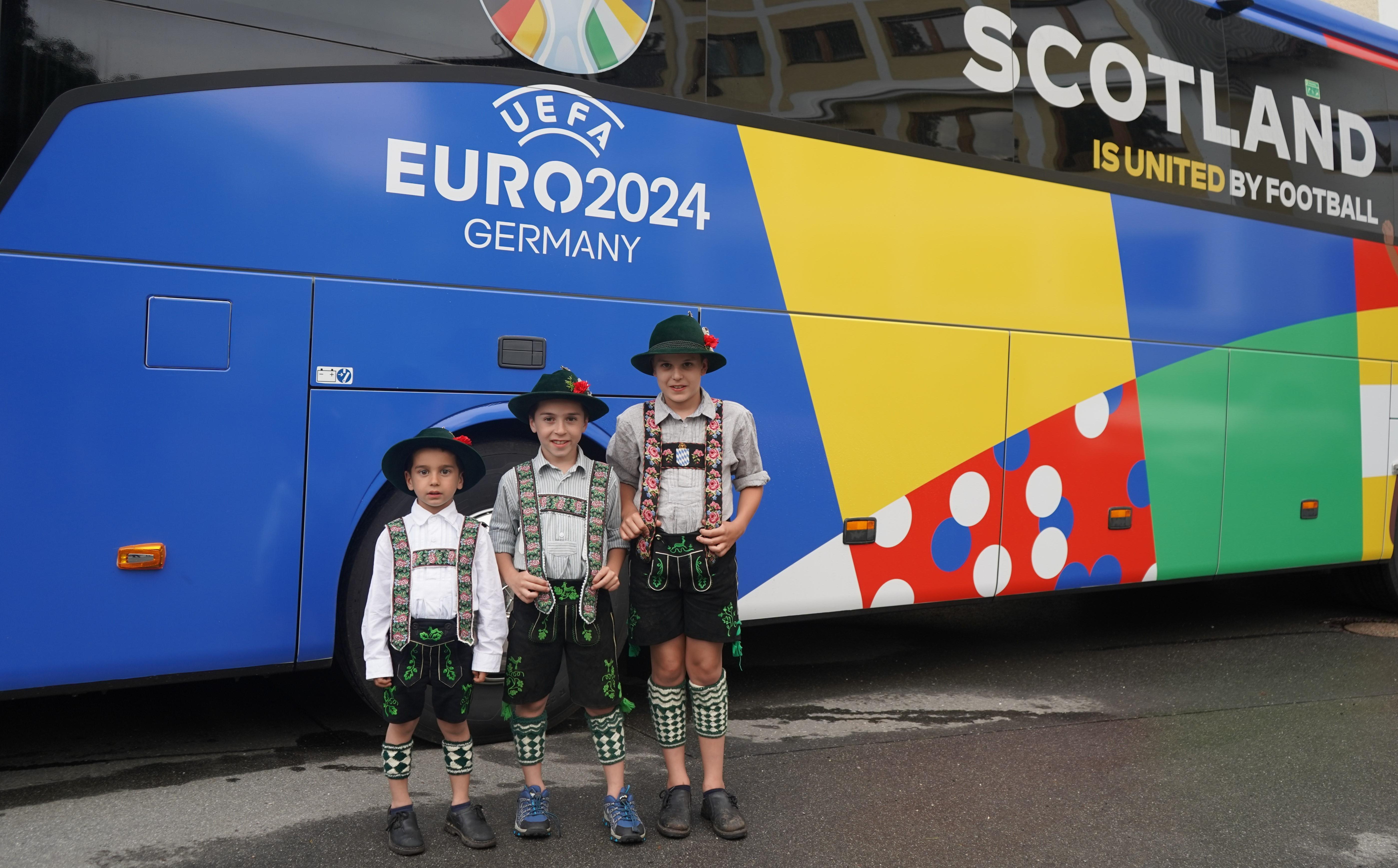 Drei Jungen in bayerischer Tracht stehen vor dem schottischen Mannschaftsbus. 