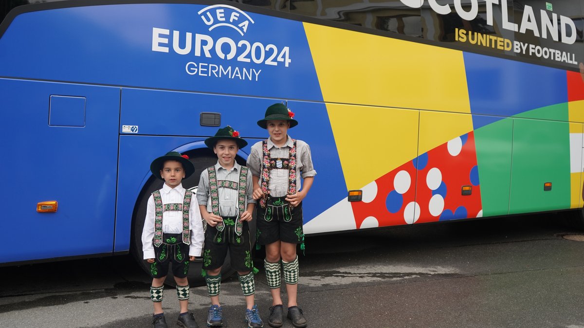 Drei Jungen in bayerischer Tracht stehen vor dem schottischen Mannschaftsbus. 