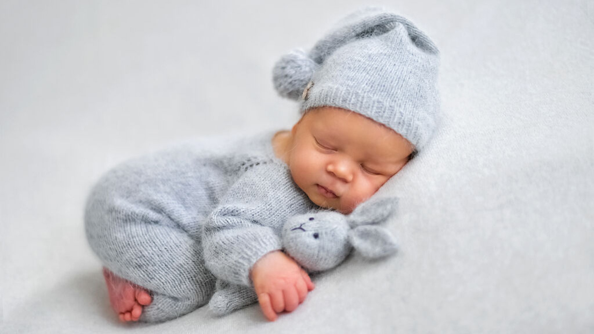 Ein Baby schläft mit einem Kuscheltier im Arm auf einer grauen Decke. 