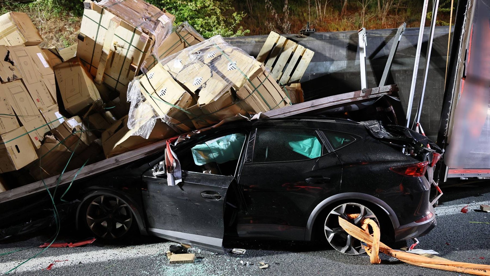 Umgestürzter Lkw auf der Autobahn, Kartons und Paletten liegen verstreut. Ein beschädigtes Auto ist unter der Ladung eingeklemmt. Einsatzkräfte sichern die nächtliche Unfallstelle.