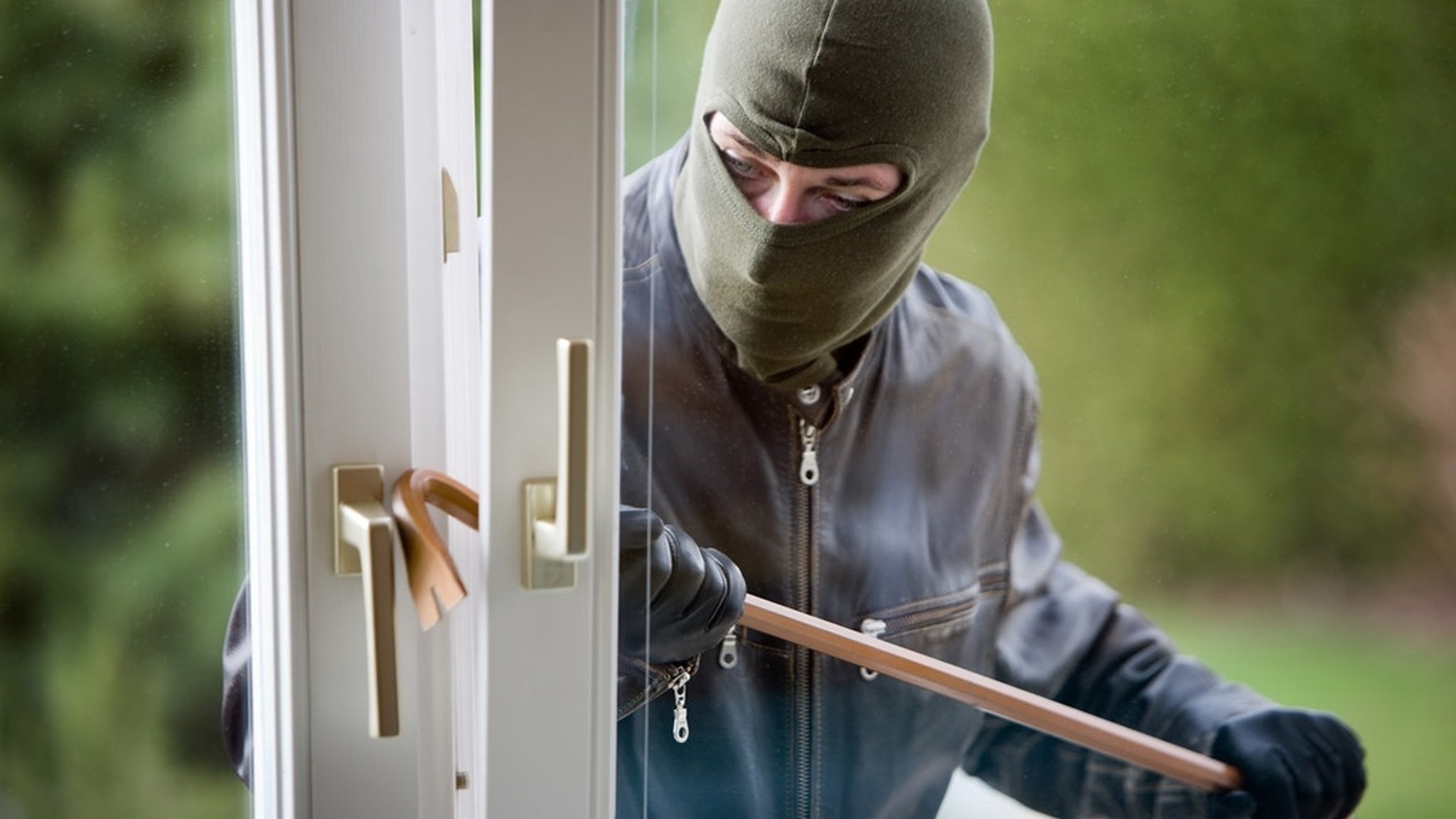 Ein vermummter Mann hebelt ein Fenster mit einem Brecheisen auf (Symbolbild).