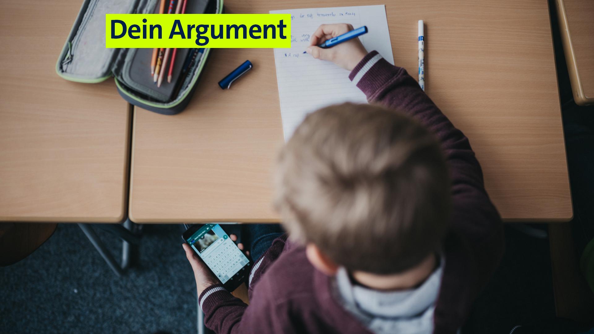 Ein Schüler blickt auf sein Smartphone, das er unter dem Tisch in seiner Hand hält. Währenddessen schreibt er mit einem Füller auf ein Blatt Papier. 