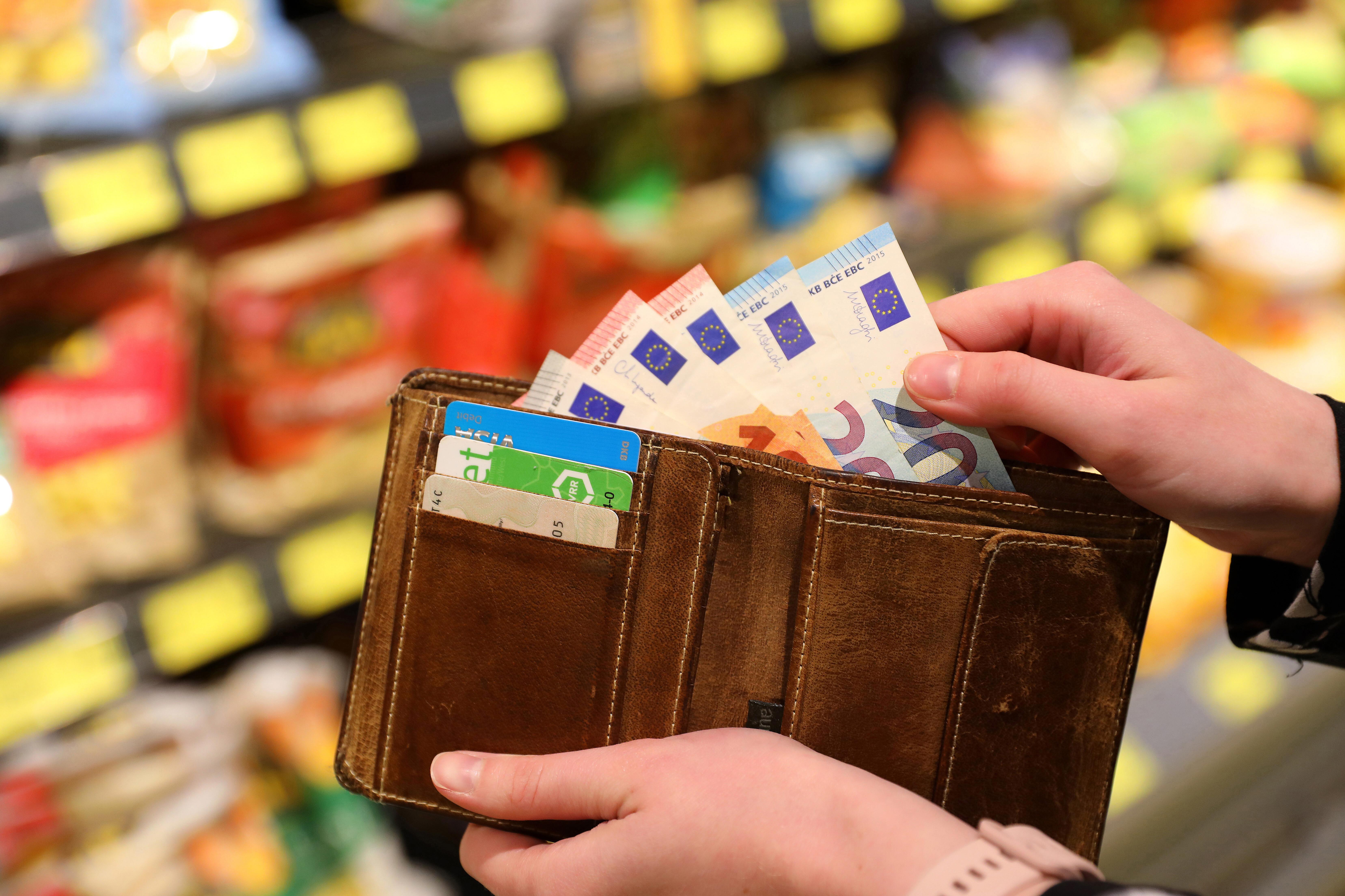 Symbolbild: Hände halten Geldbeutel mit Euro-Scheinen, im Hintergrund ein Supermarktregal