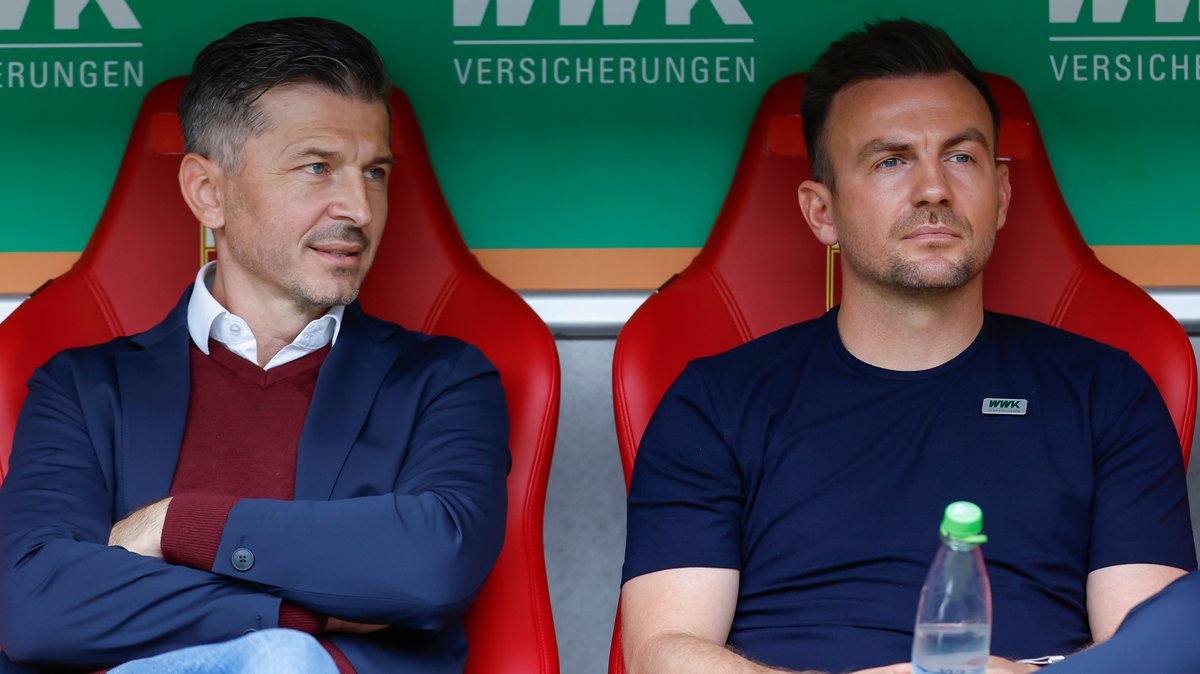 Trainer Enrico Maaßen (r) vom FC Augsburg sitzt mit skeptischem Blick auf der Spielerbank, Sportdirektor Marinko Jurendic spricht mit ihm.