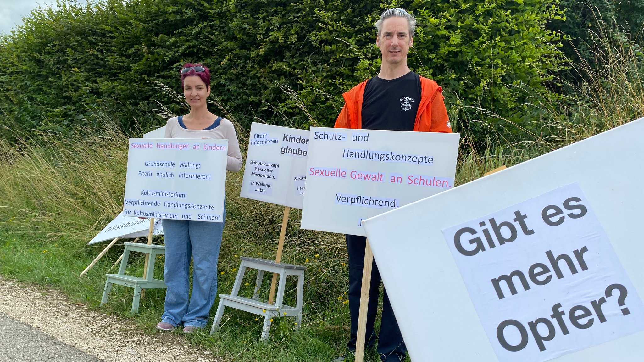 Zwei Menschen stehen mit Schildern am Straßenrand. Auf den Schildern steht etwa "Gibt es mehr Opfer?", "Schutz und Handlungskonzepte Sexuelle Gewalt an Schulen. Verpflichtend" oder "Grundschule Walting: Eltern endlich informieren!"