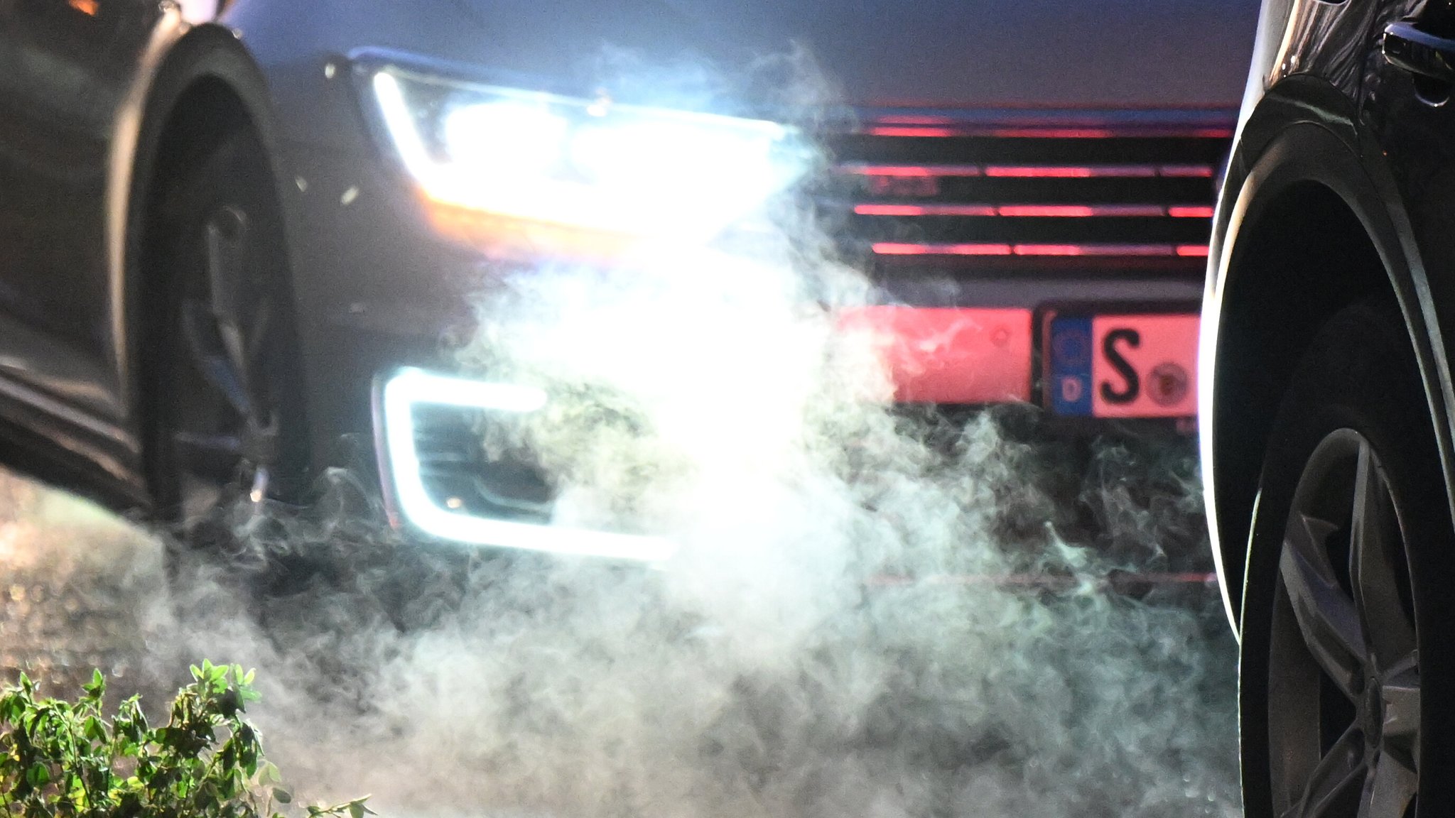 Zwei Autos stehen auf der Straße, während die Abgase vom Frontschweinwerfer des hinteren Fahrzeugs beleuchtet werden.