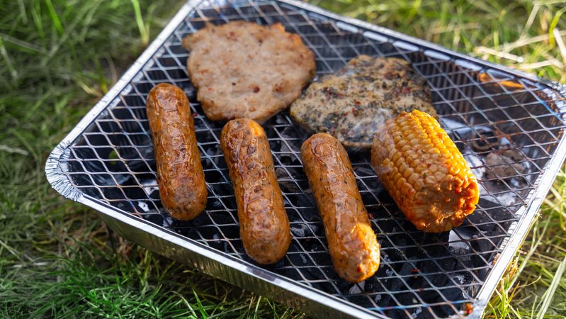 Auf einem Einweggrill liegen vegane Würstchen, ein pflanzliches Steak und ein gegrillter Maiskolben. Die Speisen braten über der Glut bei sonnigem Wetter im Freien. | Bild: picture alliance / CHROMORANGE | Michael Bihlmayer Auf einem Einweggrill liegen vegane Würstchen, ein pflanzliches Steak und ein gegrillter Maiskolben. Die Speisen braten über der Glut bei sonnigem Wetter im Freien.