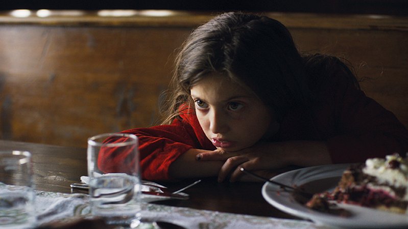 Ein kleines Mädchen sitzt vor einem Stück Torte und schaut verstört in die Kamera.