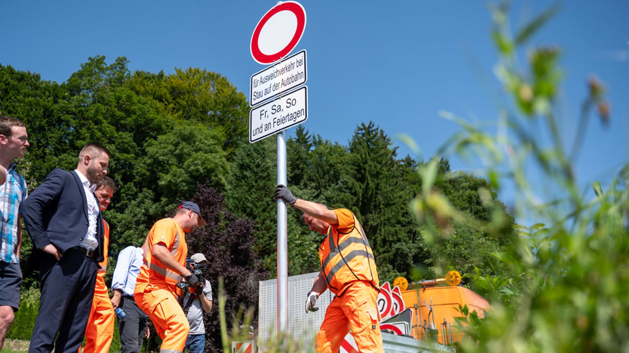 Drei Männer im orangenen Arbeitsoutfit montieren ein Verkehrsschild.  