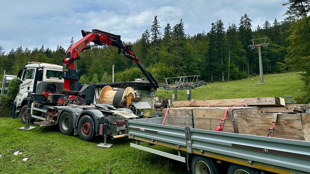 Ein Kran hebt große Holzbalken von einem Hänger am Ende einer grünen Skipiste