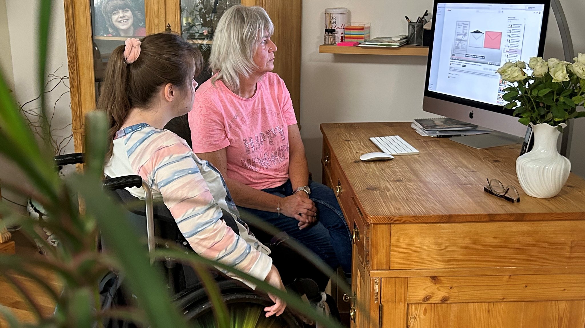 Eine junge Frau im Rollstuhl sitzt mit einer weiteren Frau an einem Schreibtisch. Sie sehen sich auf einem Monitor an, wie Breifwahl funktioniert. 