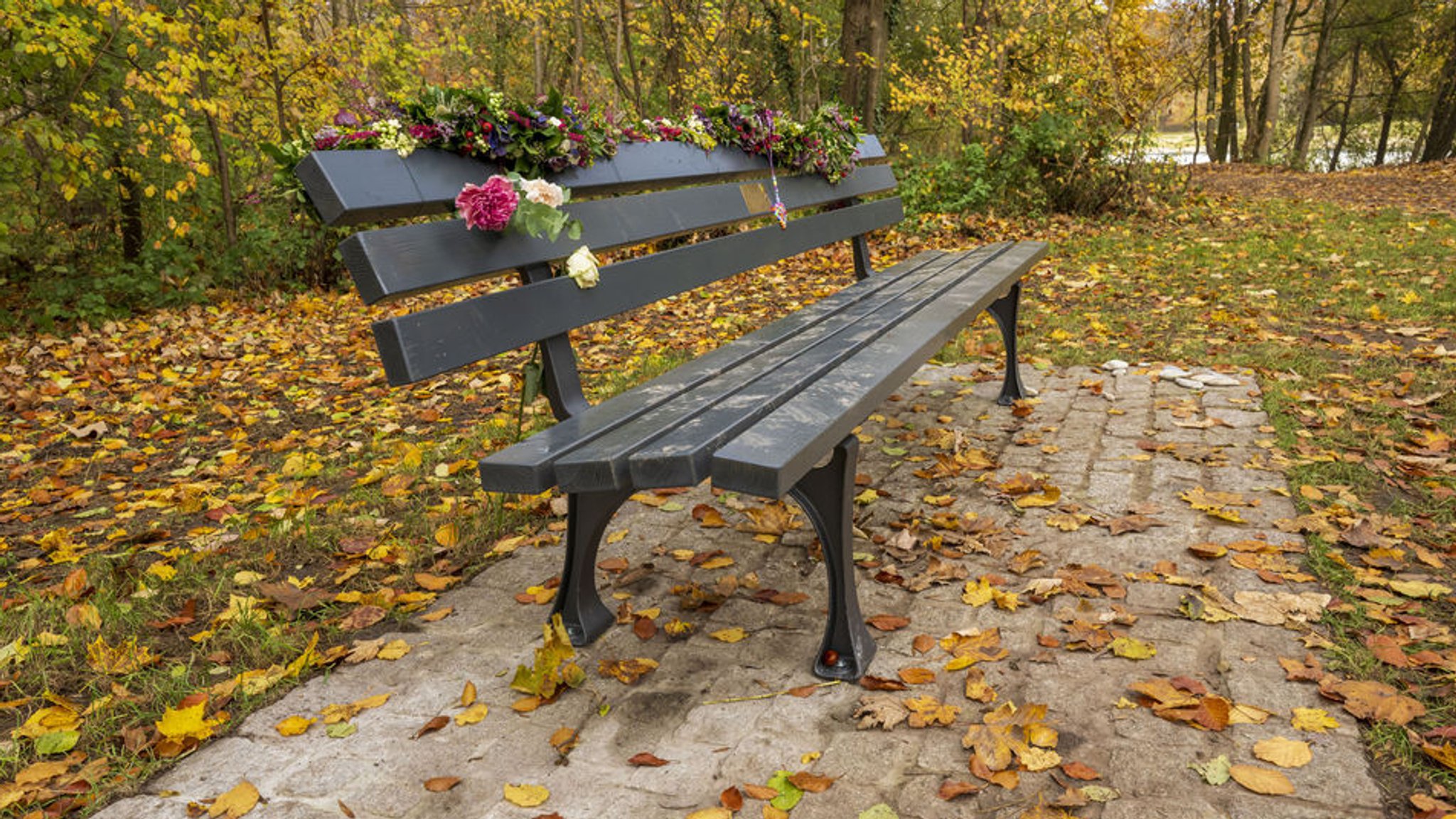 31.10.2025, Bayern, München: Eine mit Blumen geschmückte Bank steht nahe am Flaucher. Die Frau und ihre zweijährige Tochter wurden Opfer eines Anschlags, bei dem ein Mann am 13. Februar 2025 sein Auto in einen Demonstrationszug lenkte. Foto: Peter Kneffel/dpa +++ dpa-Bildfunk +++