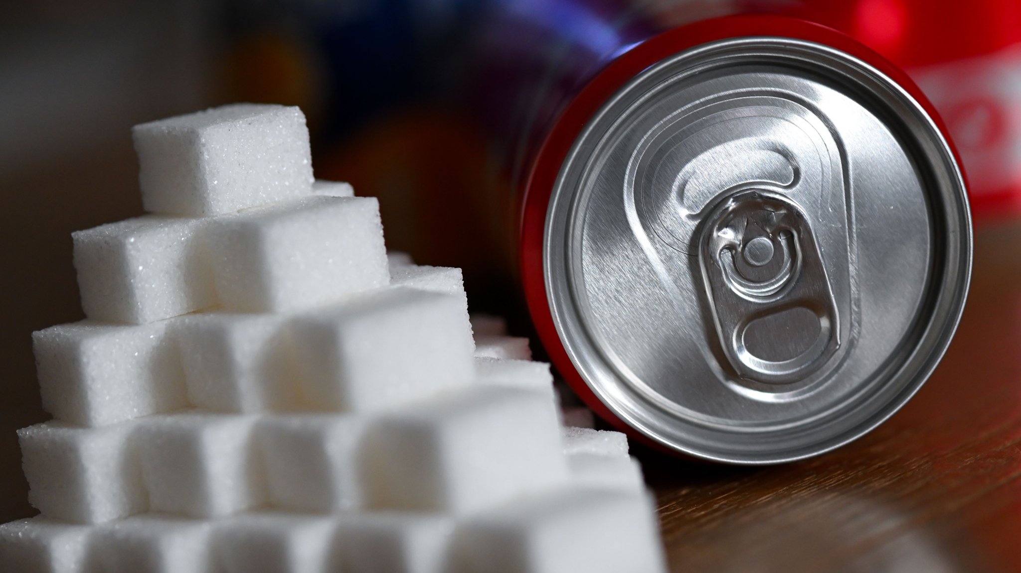 (Symbolbild) Eine Getränkedose mit einem Softdrink und zahlreiche Zuckerwürfel liegen auf einem Tisch. 