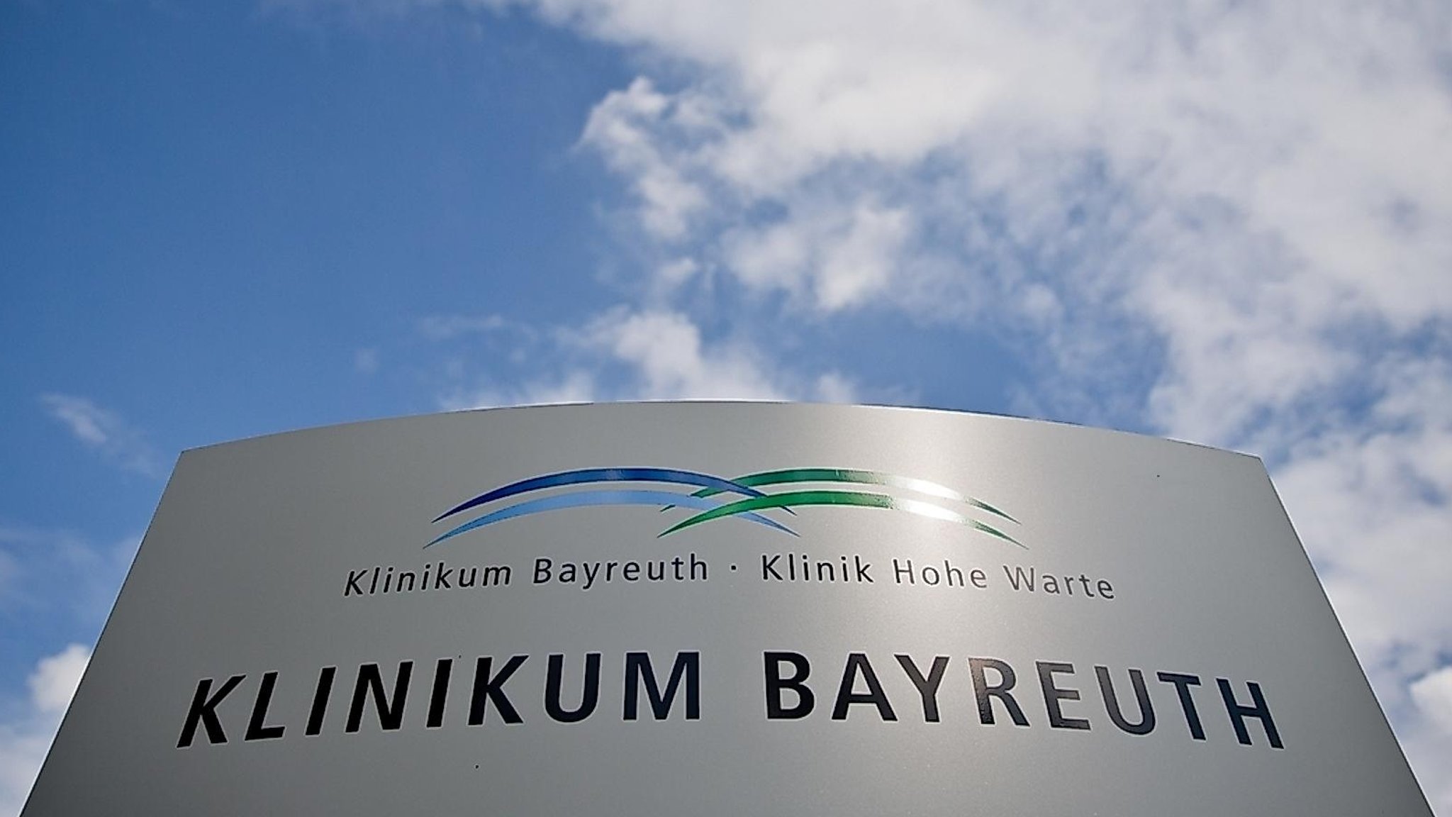 Auf einem Schild ist zu lesen: Klinikum Bayreuth, Klinik Hohe Warte, im Hintergrund weiß-blauer Himmel.