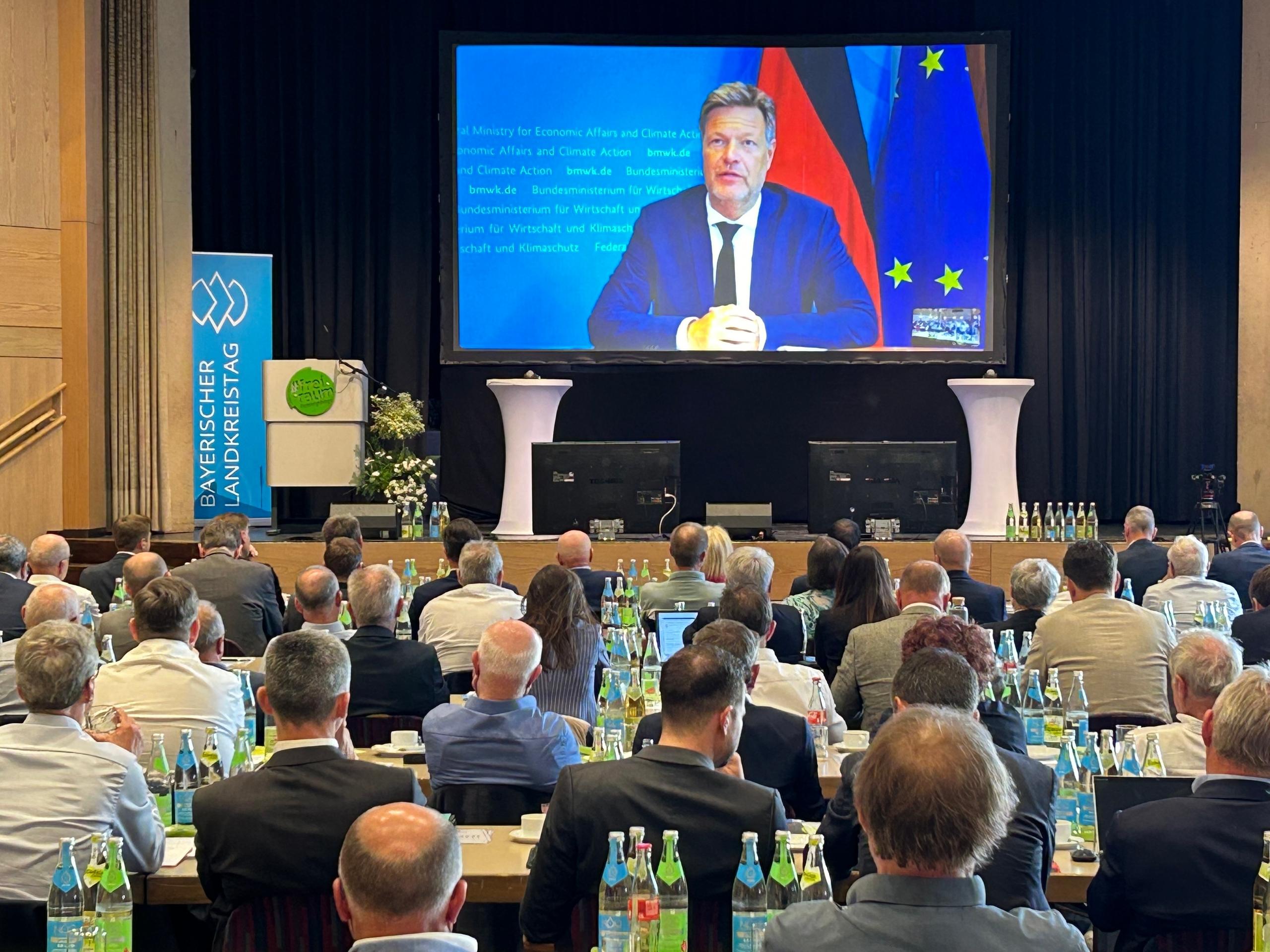 In einem Saal sitzen viele Männer und schauen auf Bundeswirtschaftsminister Robert Habeck, der per Video zugeschaltet  ist. 