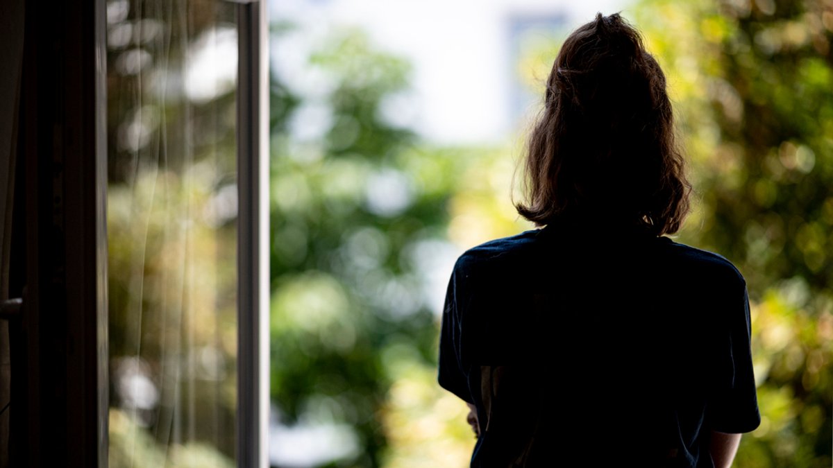 Eine junge Frau steht in ihrer Wohnung an einem Fenster.