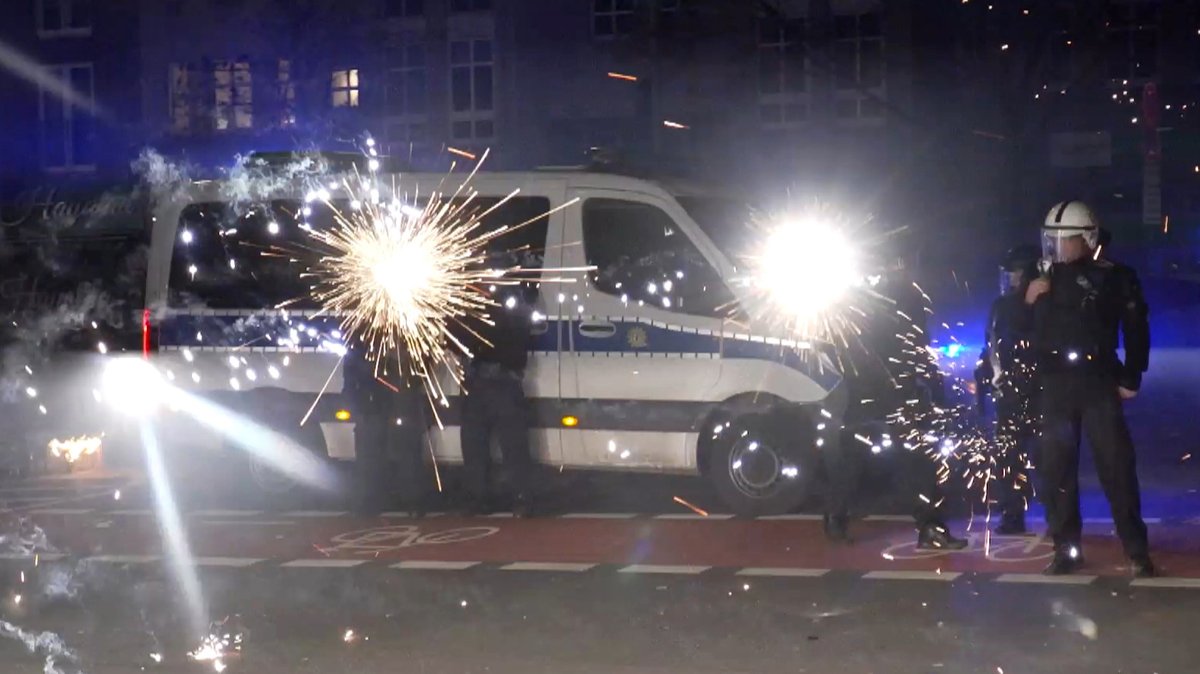 Polizisten stehen an einem Einsatzwagen, um sie herum explodieren Feuerwerkskörper.