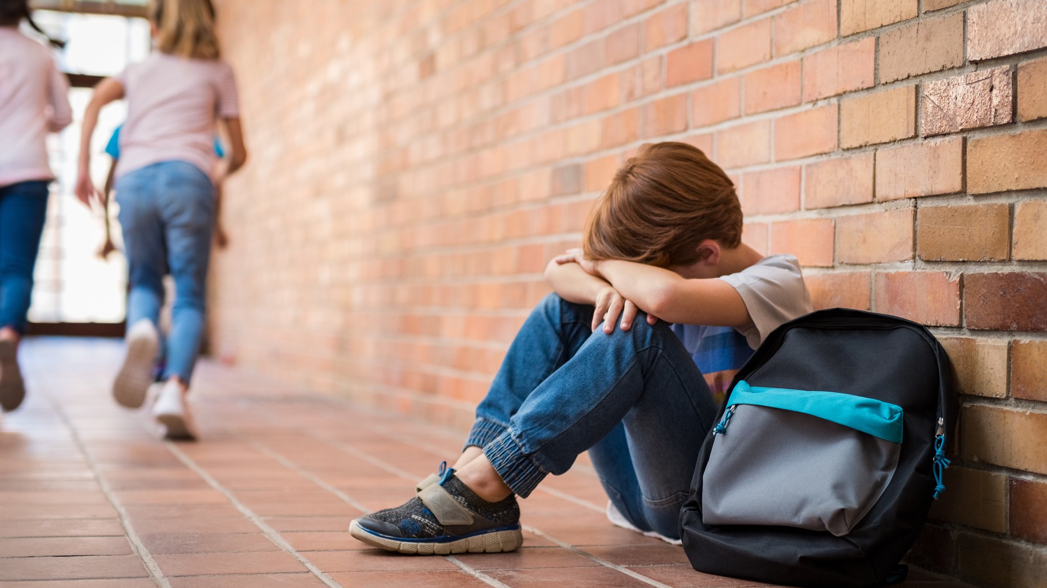 Symbolbild. Ein Junge sitzt auf dem Flur in seiner Schule mit dem Rücken zur Wand und legt seinen Kopf zwischen seine Arme. Neben ihm liegt sein Schulranzen, Kinder laufen durch den Flur nach draußen. | Bild: stock.adobe.com/Rido Symbolbild. Ein Junge sitzt auf dem Flur in seiner Schule mit dem Rücken zur Wand und legt seinen Kopf zwischen seine Arme. Neben ihm liegt sein Schulranzen, Kinder laufen durch den Flur nach draußen.