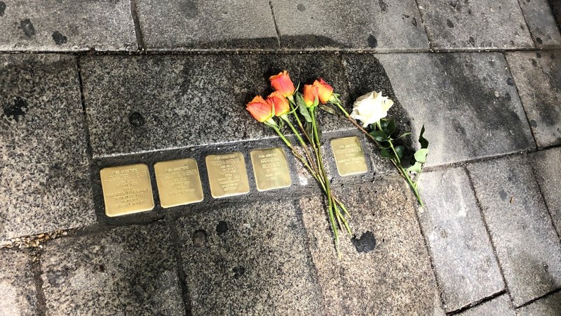 Stolpersteine, in den Boden eingelassene, vergoldete Steine mit Daten verfolgter und ermordeter Opfer des Nationalsozialismus. Neben den Steinen liegen mehrere rot-gelbe Rosen und eine weiße Rose.