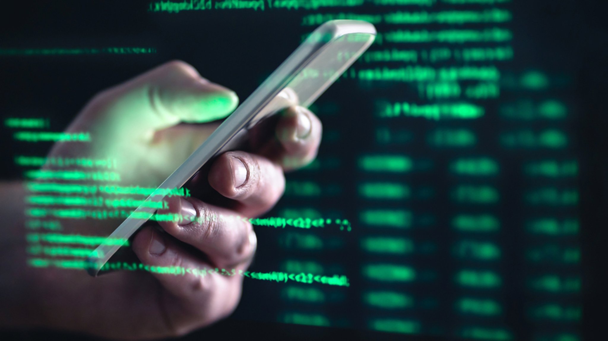 Eine Hand mit Smartphone spiegelt sich in einem Bildschirm mit grünen Computercodes