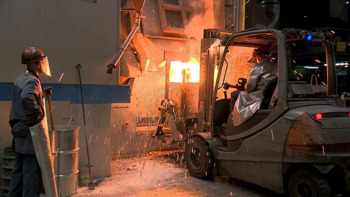 Stahlarbeiter in Schutzkleidung stehen vor einem Gabelstapler, im Hintergrund sprühen Funken. 