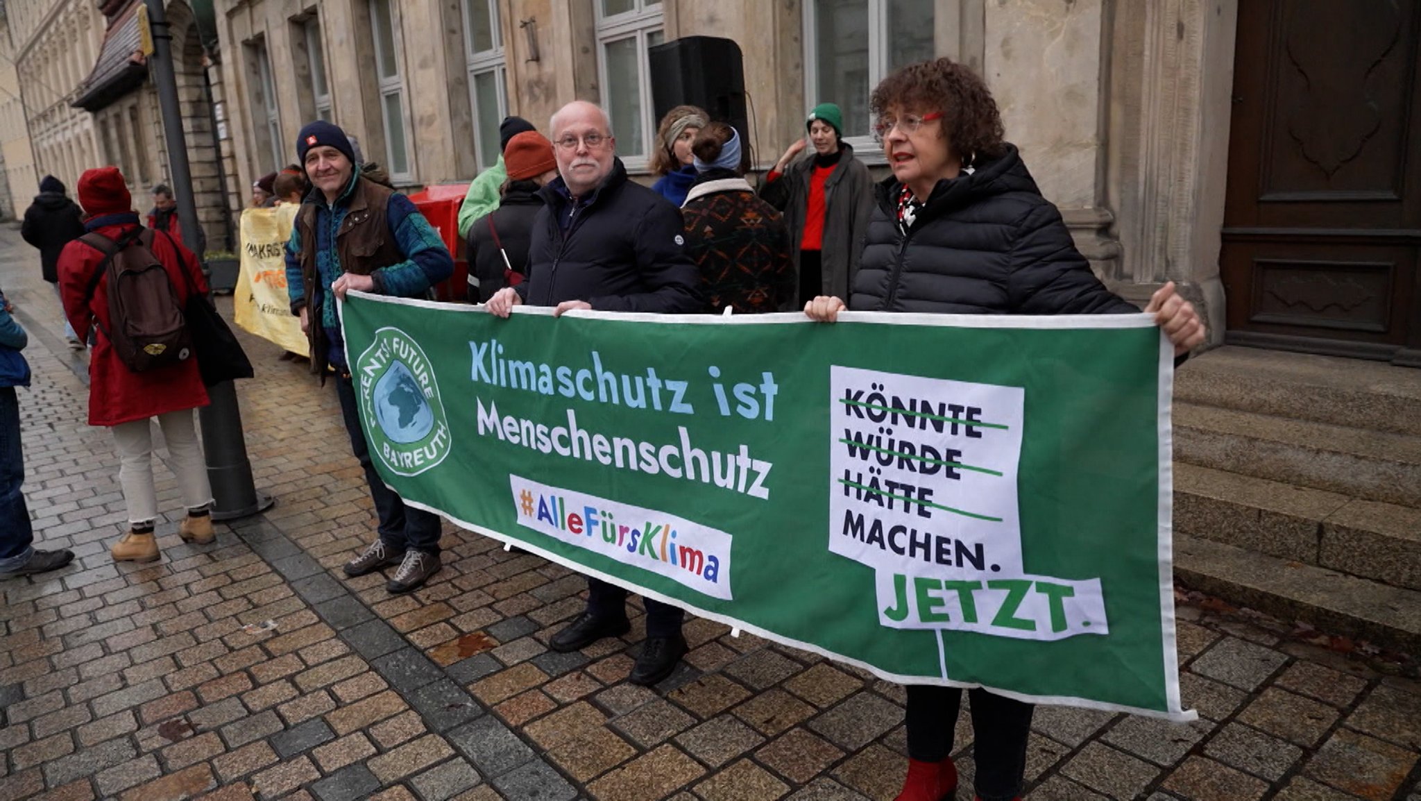 Vor einem Gerichtsgebäude stehen Menschen und halten Transparente, auf denen unter anderem steht:" Klimaschutz ist Menschenschutz". 