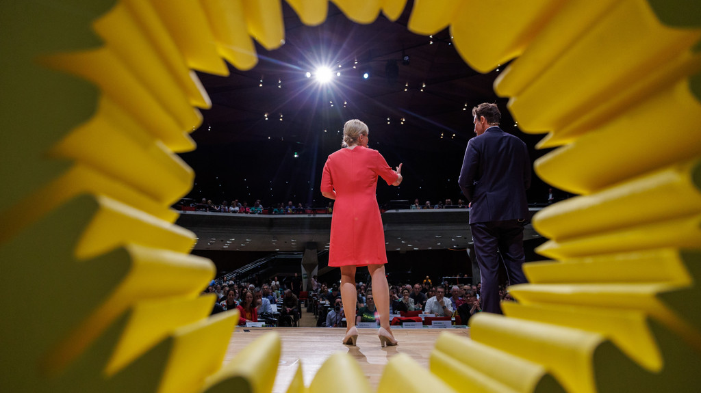 Katharina Schulze und Ludwig Hartmann, Spitzenkandidaten der bayerischen Grünen für die Landtagswahl, auf der Bühne während des Parteitags. Fotografiert von hinten durch ein aufgestelltes Sonnenblumen-Symbol.